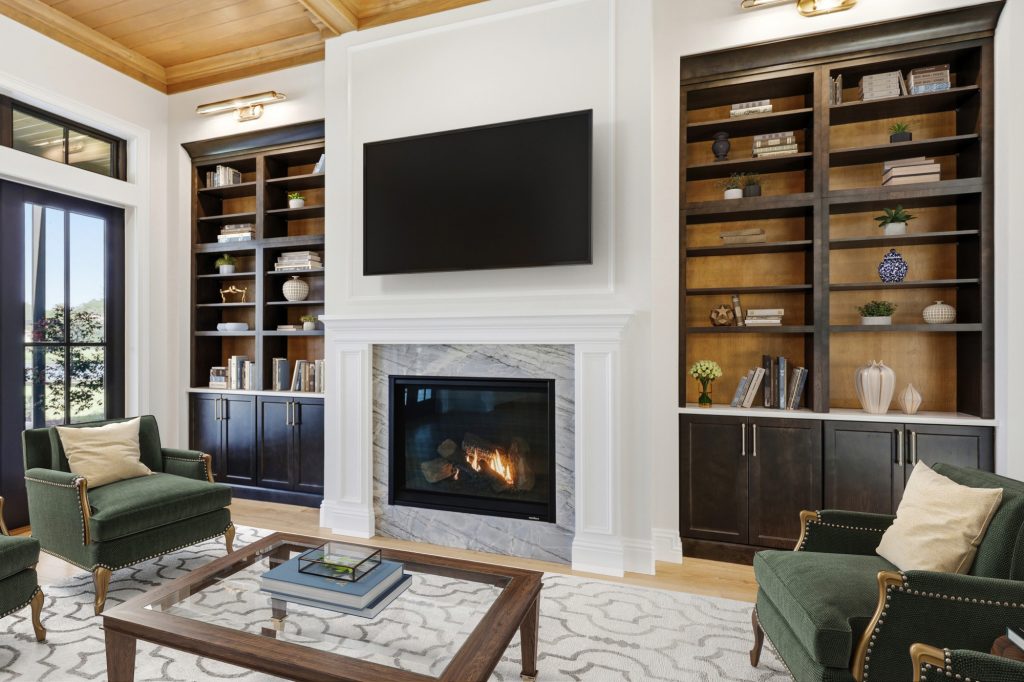 Living room with a marble-surround fireplace, dark built-in bookcases, green armchairs, and a wood-paneled ceiling.